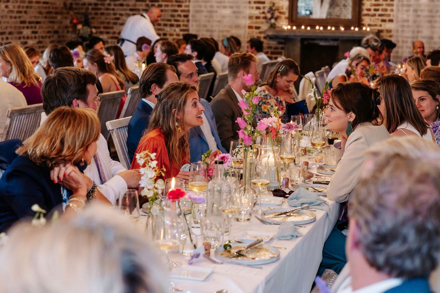 Wedding guests seated at long decorated dinner table with candles and floral centerpieces in brick venue.