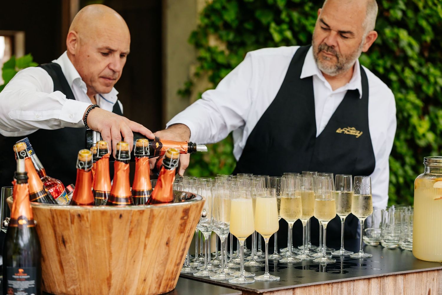 Two bartenders preparing champagne flutes and bottled drinks at a wedding reception bar.