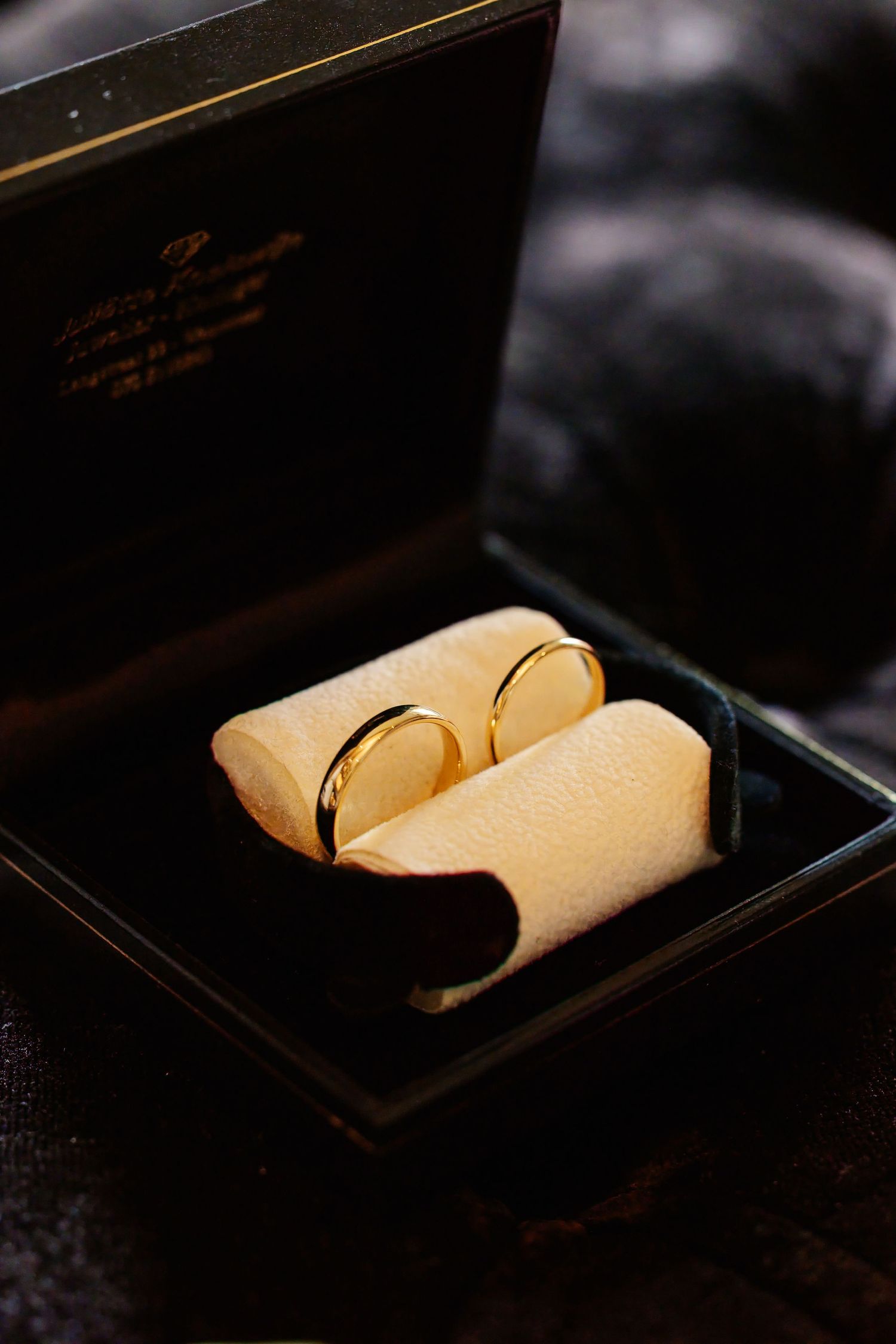 Two gold wedding bands displayed in a black velvet jewelry box with cream foam ring holders.