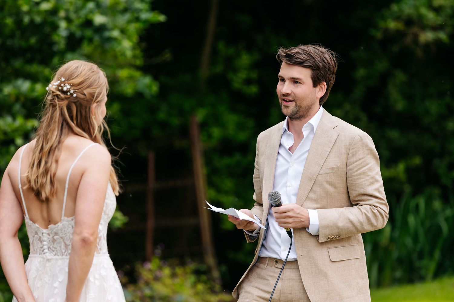 Groom in beige suit reading wedding vows to bride outdoors with lush green trees in background.