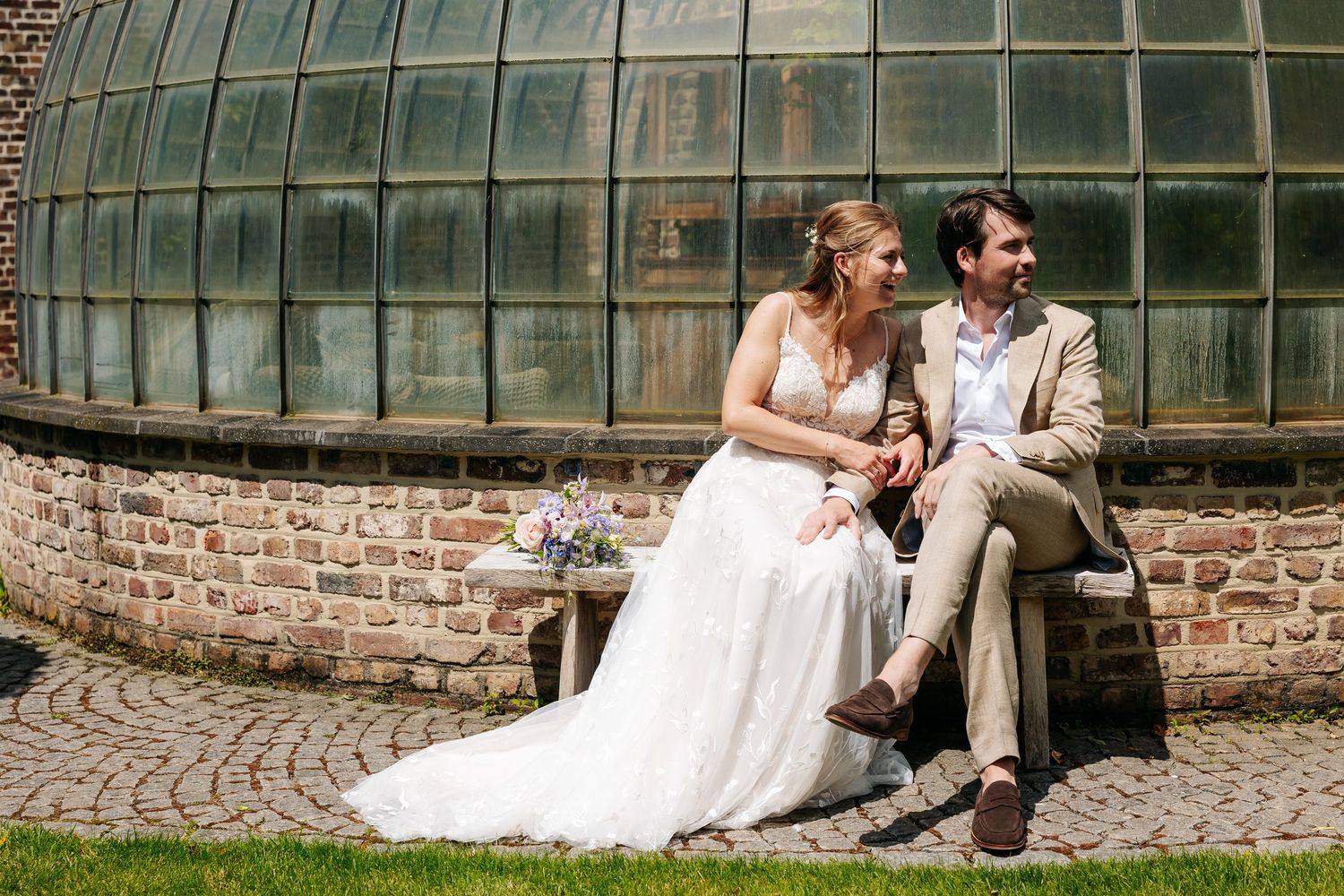 Bride and groom sitting together laughing beside a brick greenhouse wall on their wedding day.
