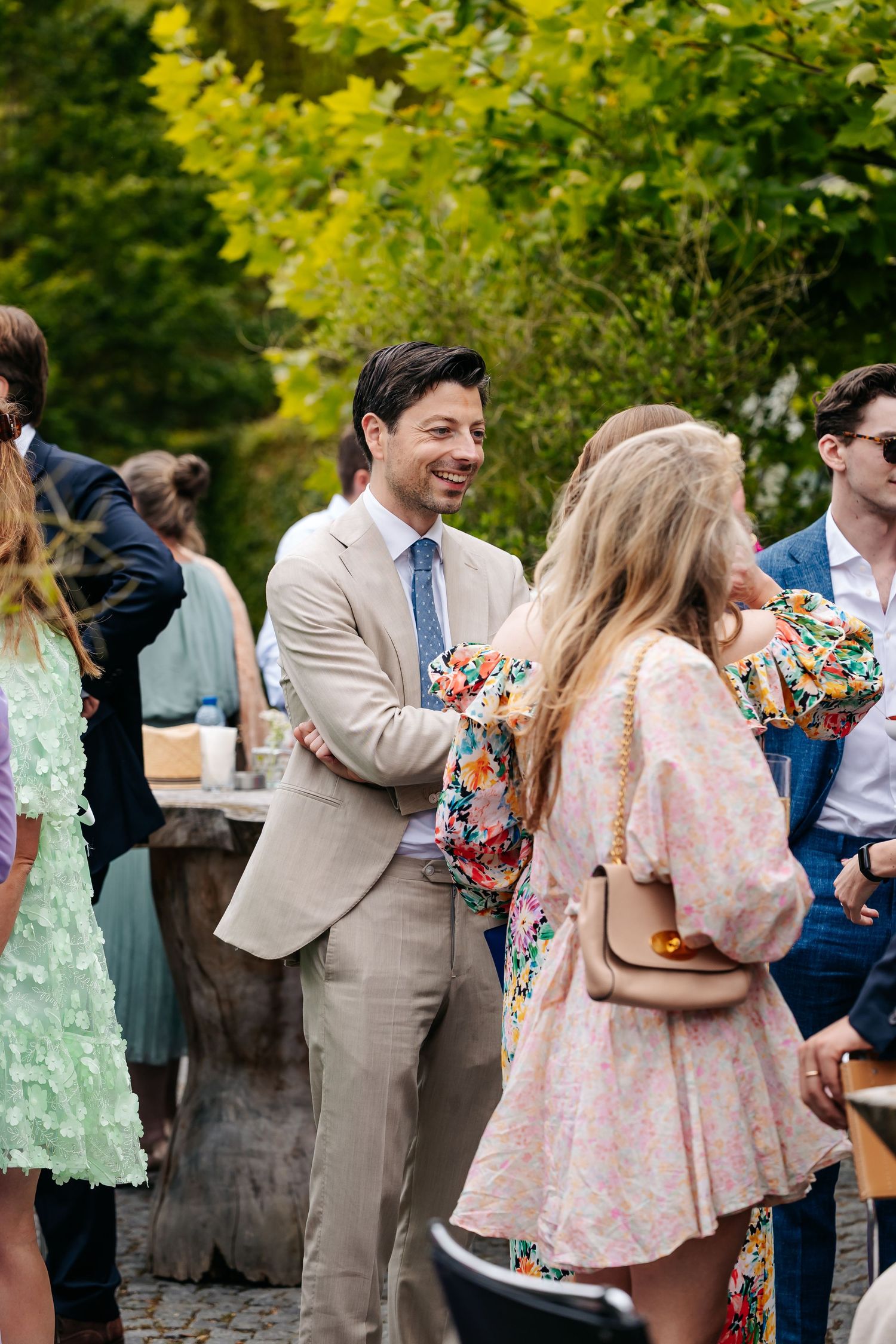 Smiling man in beige suit chats with guests in floral dresses at an outdoor garden wedding reception.