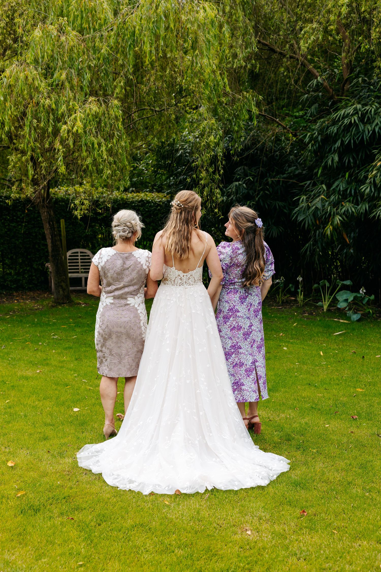 Bride in white lace gown with two women in garden, viewed from behind.