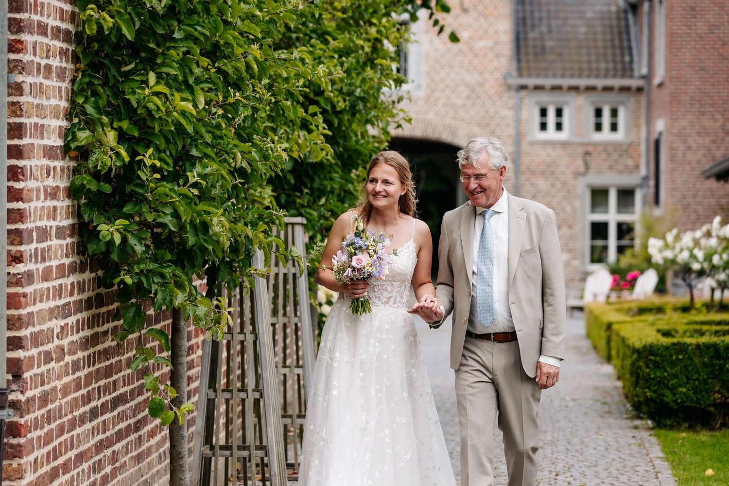 Bride in a lace wedding gown walking with her father along a brick garden path.