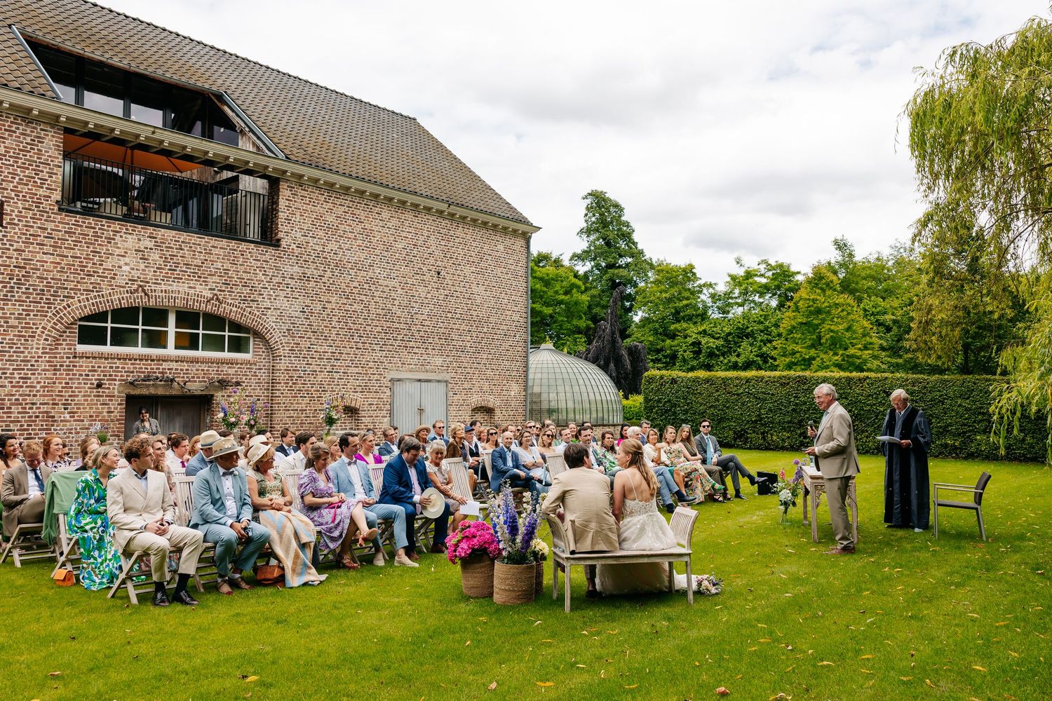 Outdoor wedding ceremony with seated guests on a sunny lawn beside a rustic brick building.