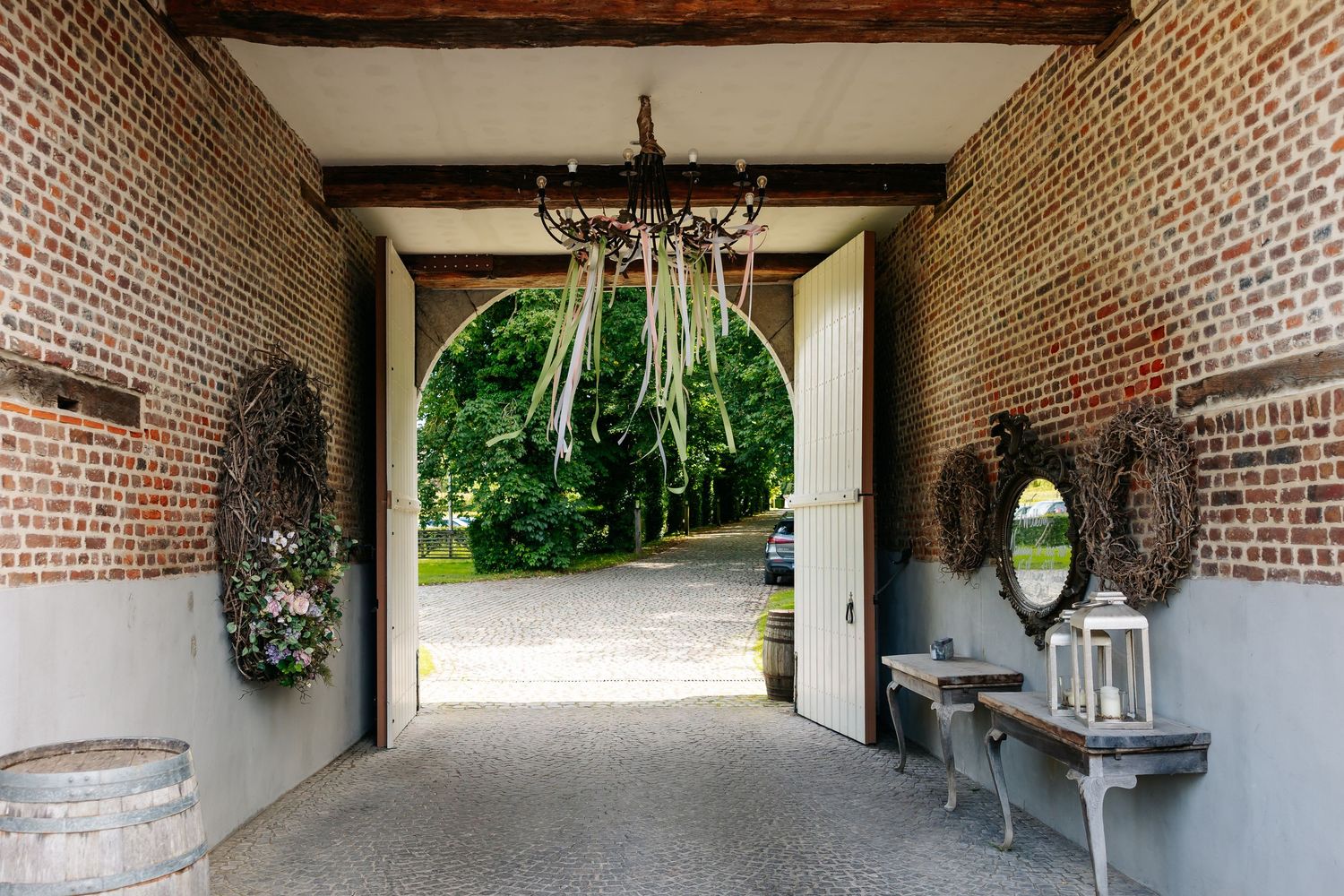 Rustic brick archway entrance with chandelier, wreath decor, and open doors revealing a lush green driveway beyond.