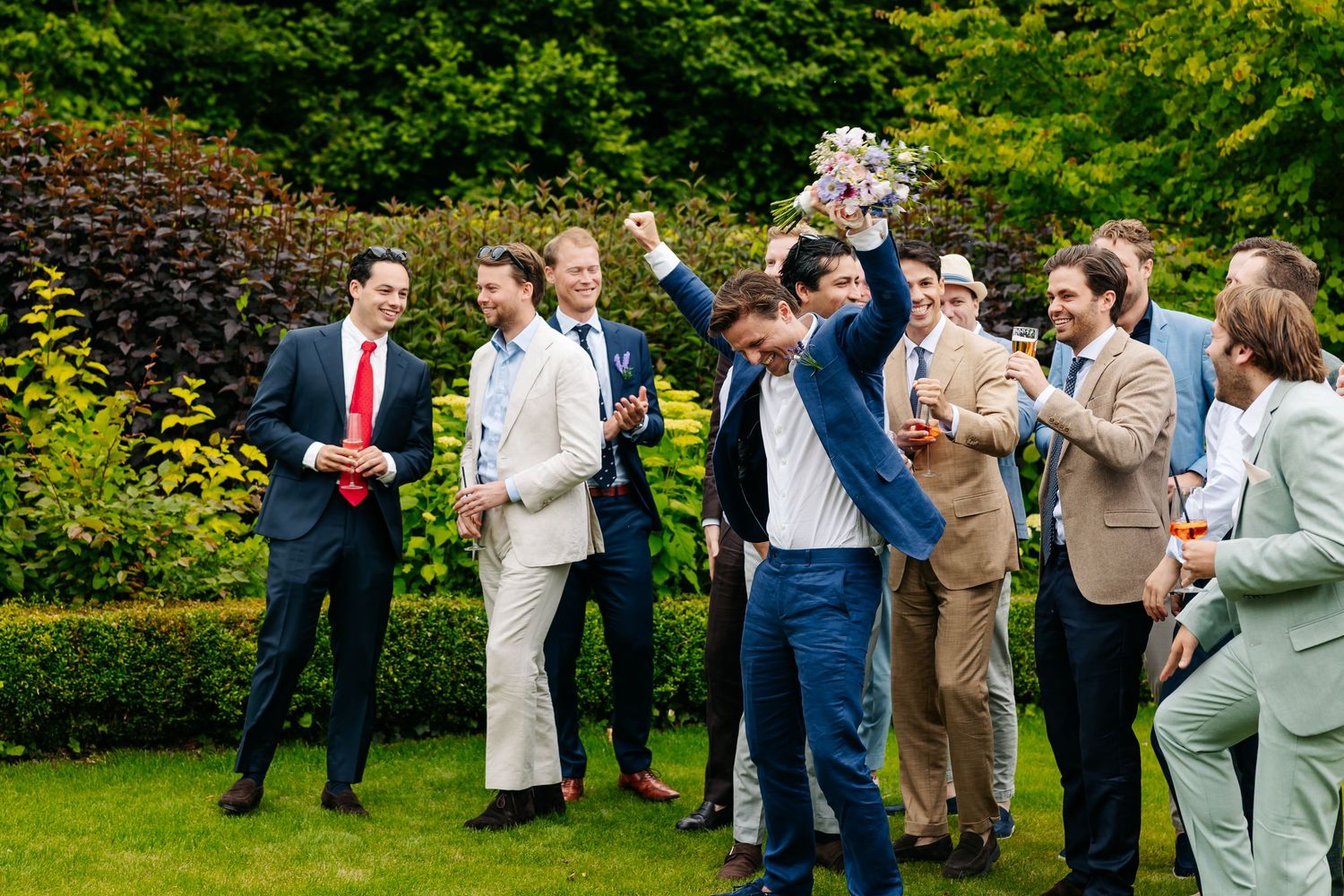 Groomsmen cheering and catching the wedding bouquet toss on a sunny garden lawn.