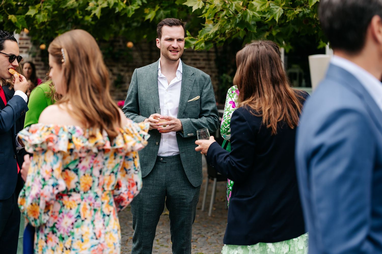 A man in a green suit smiles and chats with guests at an outdoor wedding reception.