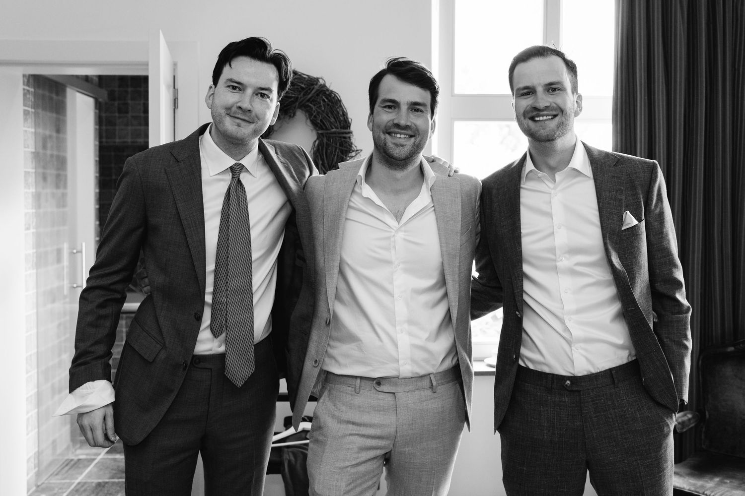 Three well-dressed men in suits smiling together in a black and white wedding photo.