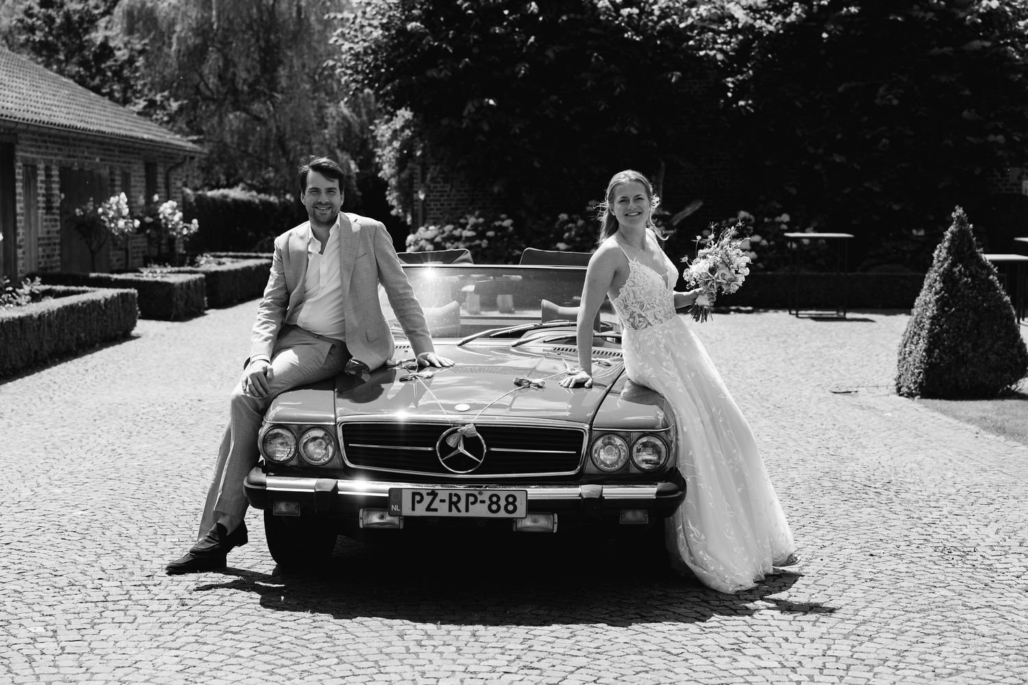 Black and white photo of bride and groom posing on vintage Mercedes-Benz convertible.