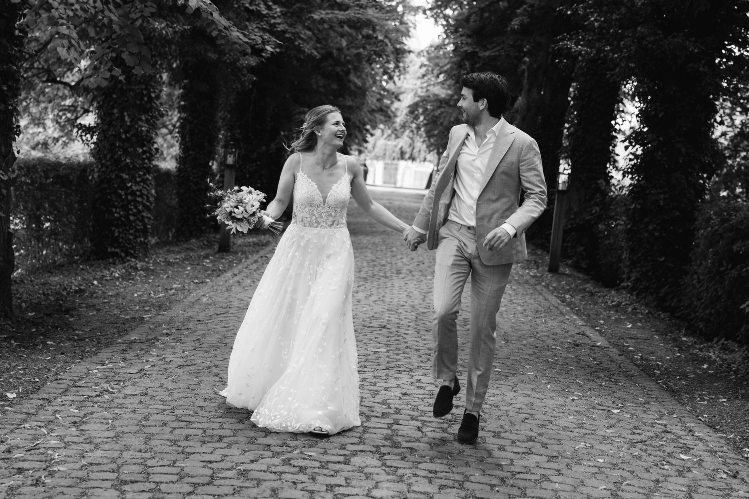 Black and white photo of smiling bride and groom walking together on tree-lined cobblestone path.