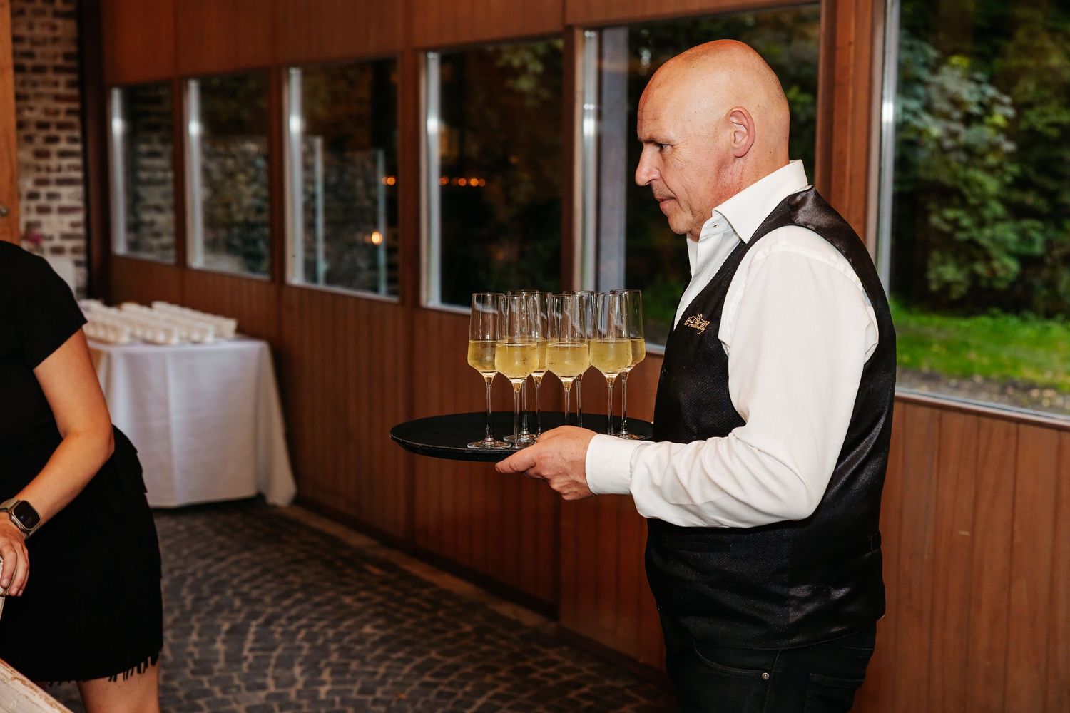 A waiter in a black vest carries a tray of champagne glasses at a wedding reception.