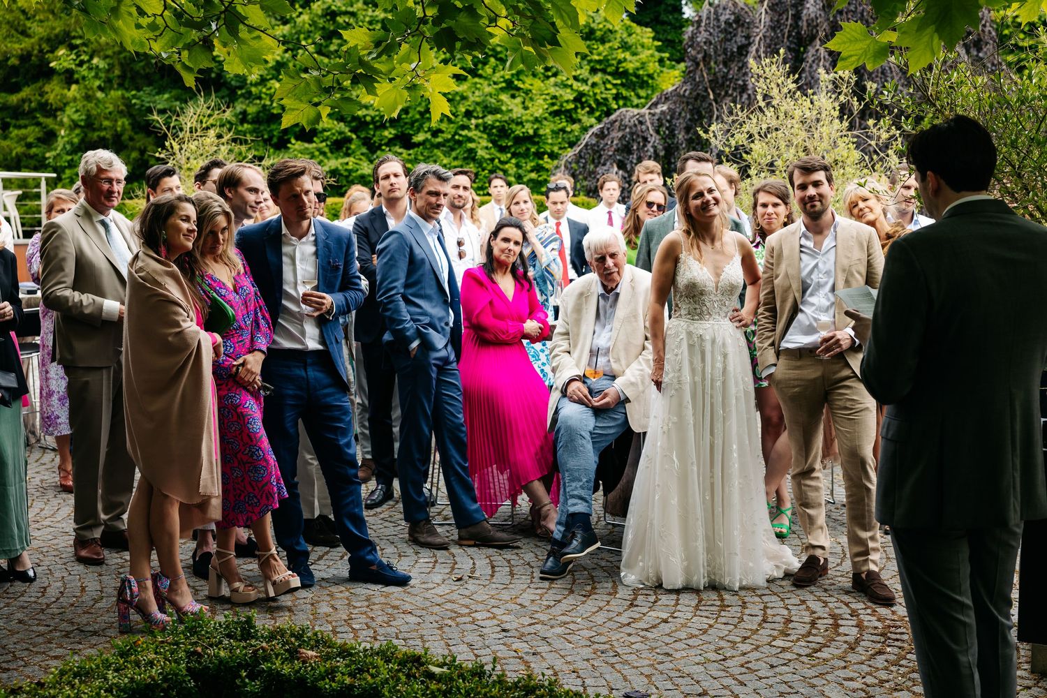 Wedding guests gathered outdoors on cobblestone patio watching bride in white gown during reception.