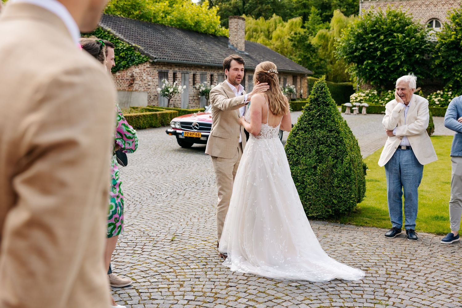 Groom in beige suit greeting bride in white wedding gown on a cobblestone driveway outdoors.