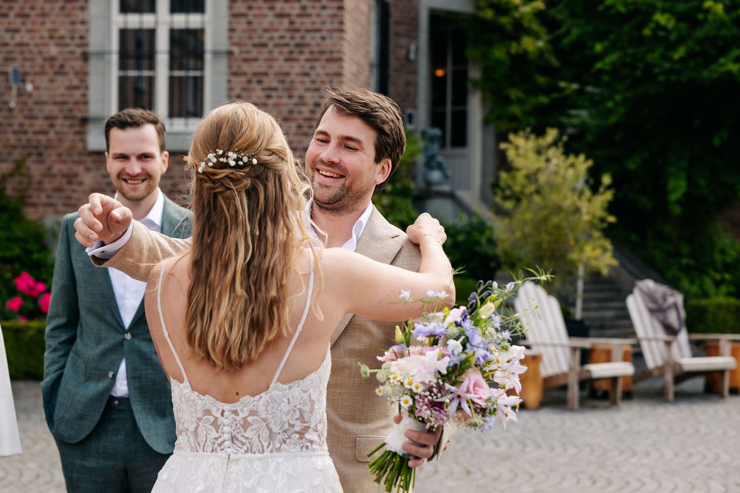 Groom in beige suit embracing bride in white lace dress holding colorful bouquet at outdoor wedding.