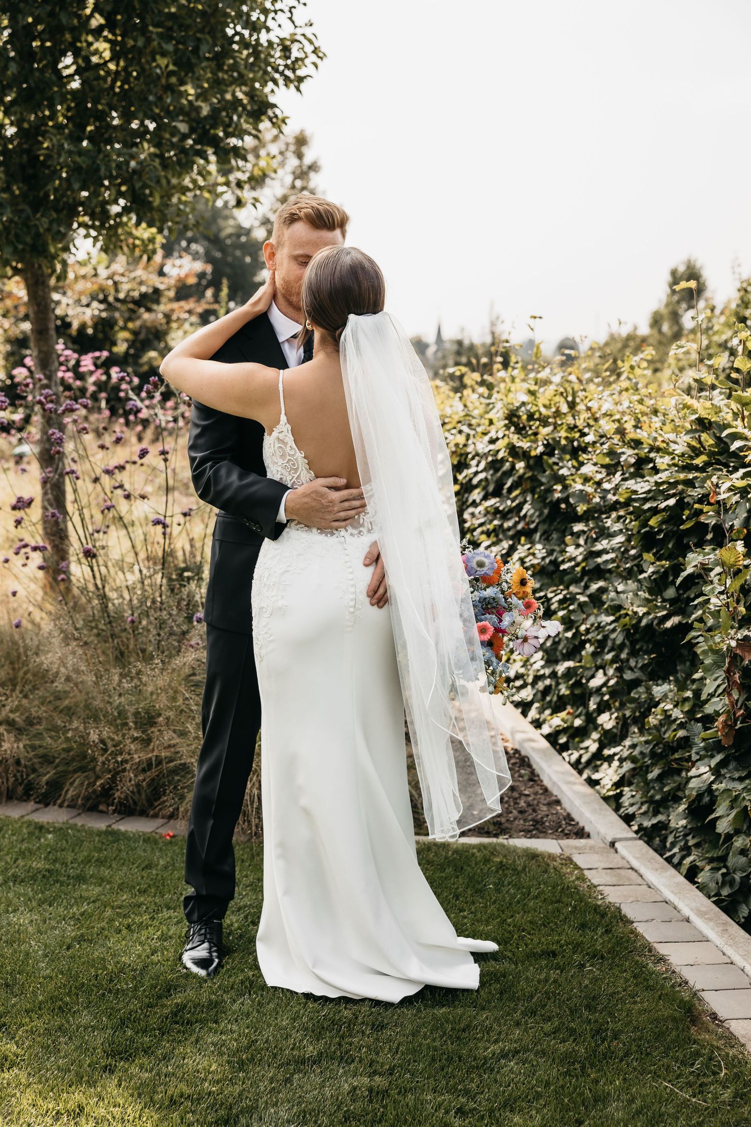 Groom embracing bride from behind in a garden, bride wearing a stunning backless lace wedding gown.