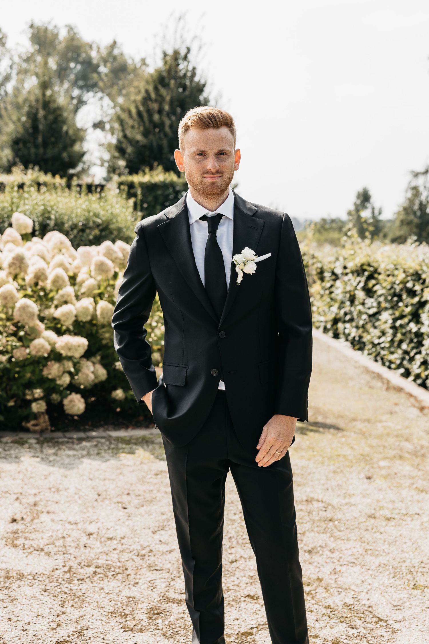 Elegant groom in black suit and tie with white boutonniere posing in a lush garden.