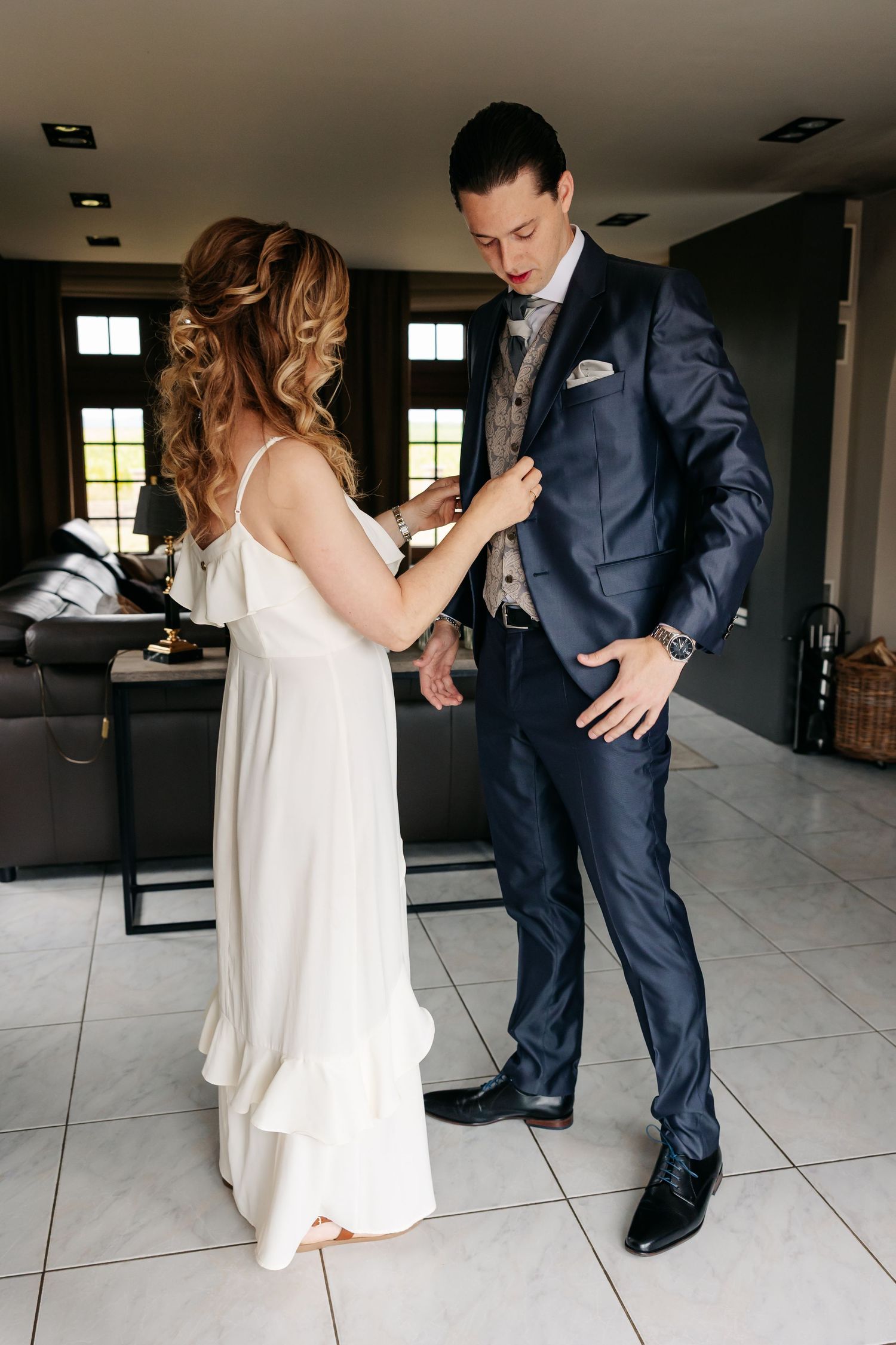 Bride in white dress pinning boutonniere on groom in navy suit before wedding.