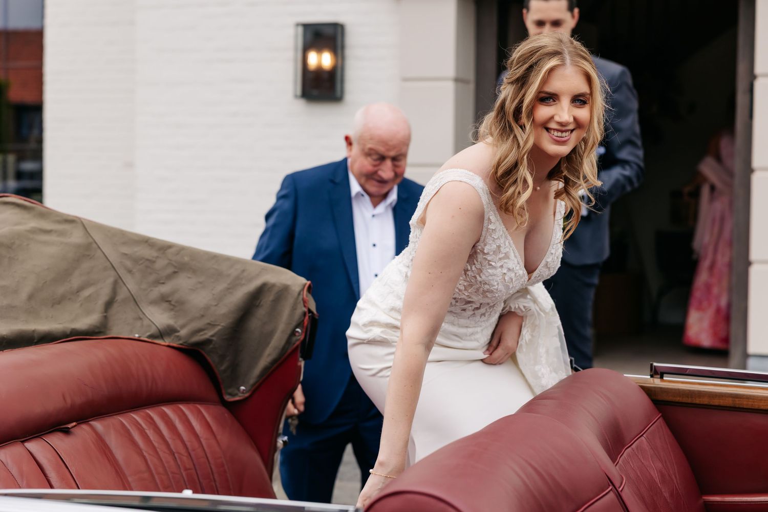 Smiling bride in lace gown climbs into classic vintage convertible car with red leather interior at wedding venue.