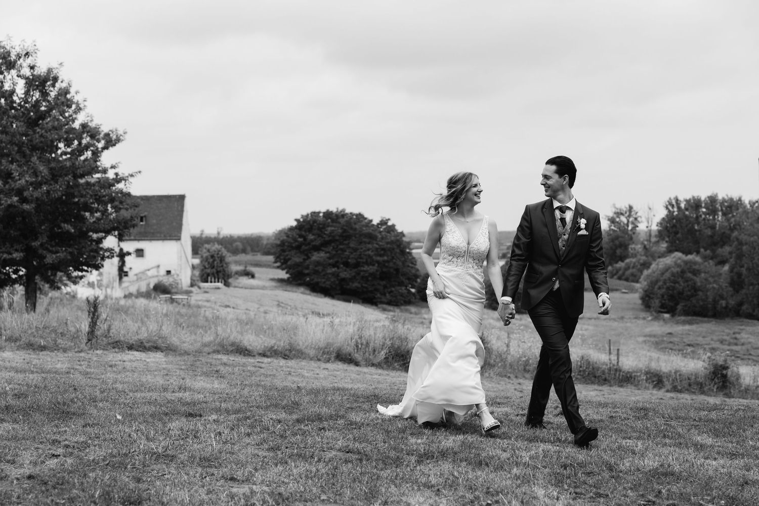 Black and white photo of newlyweds laughing and walking hand in hand across a countryside meadow.