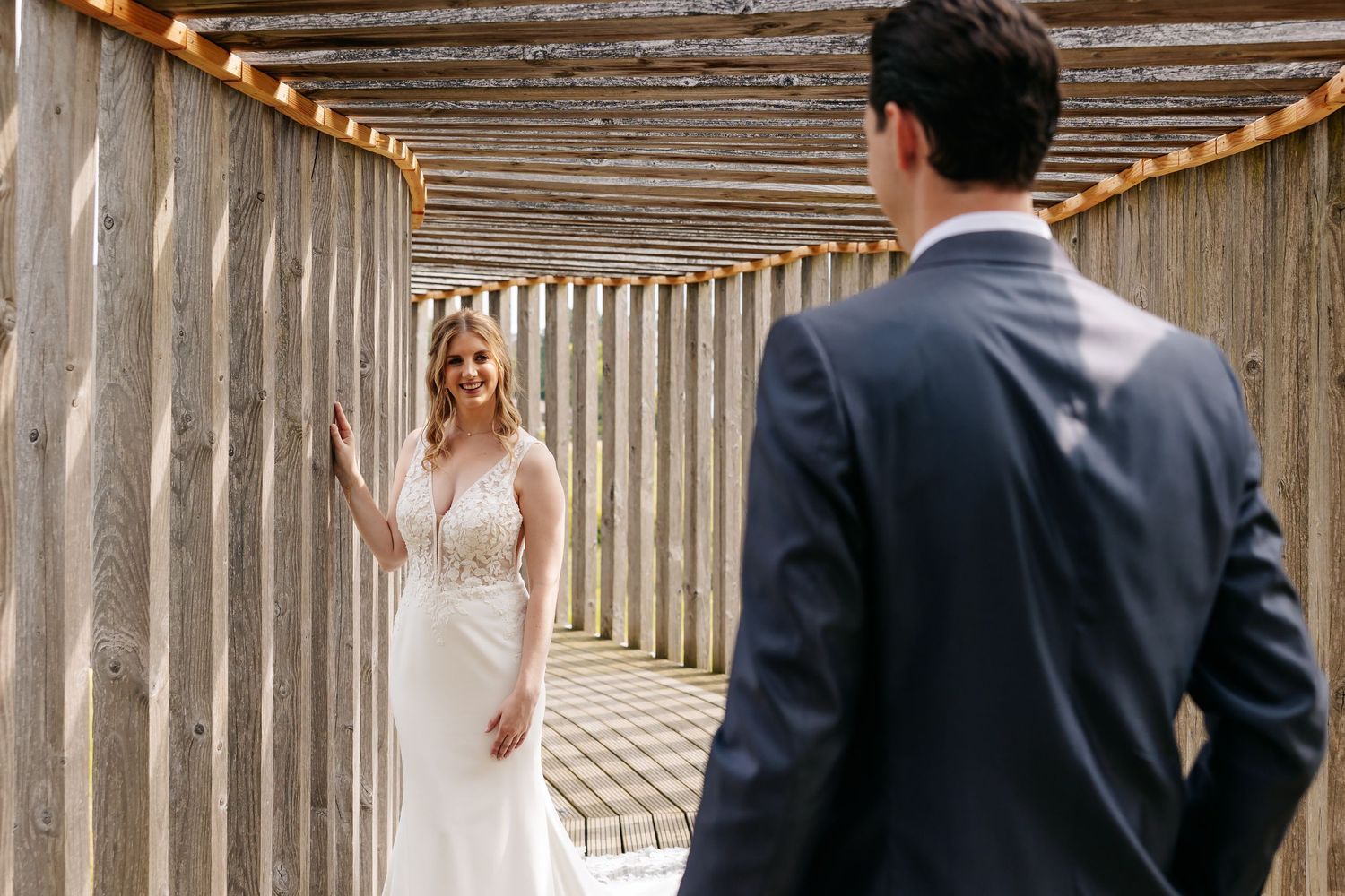 Bride in beaded wedding dress smiles at groom in wooden slatted outdoor corridor.