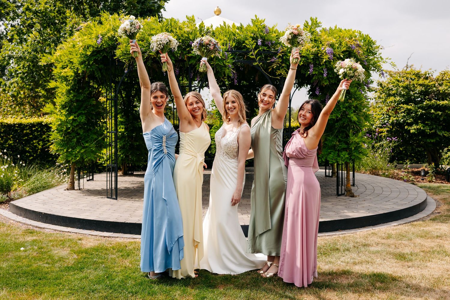 Five bridesmaids in colorful dresses raising bouquets joyfully with bride in white at outdoor wedding.