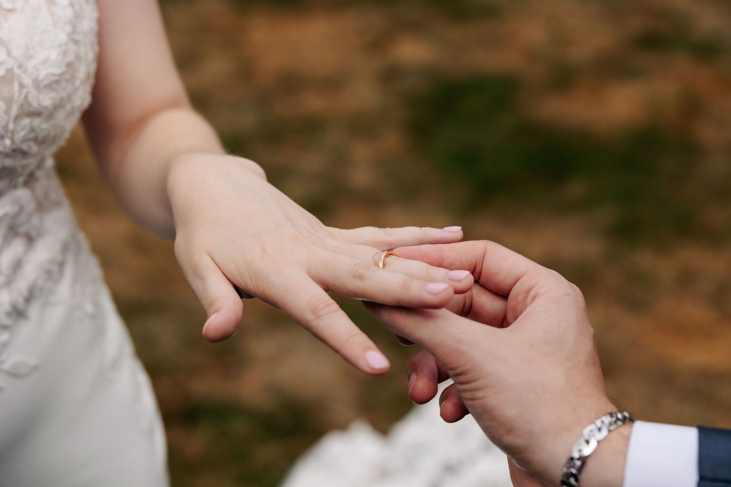 Groom placing gold wedding ring on bride's finger during outdoor wedding ceremony.