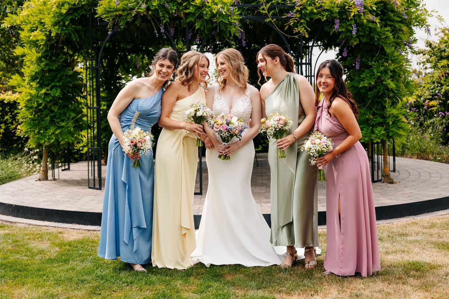 Bride in white lace gown laughs with four bridesmaids in pastel dresses holding bouquets in a garden.