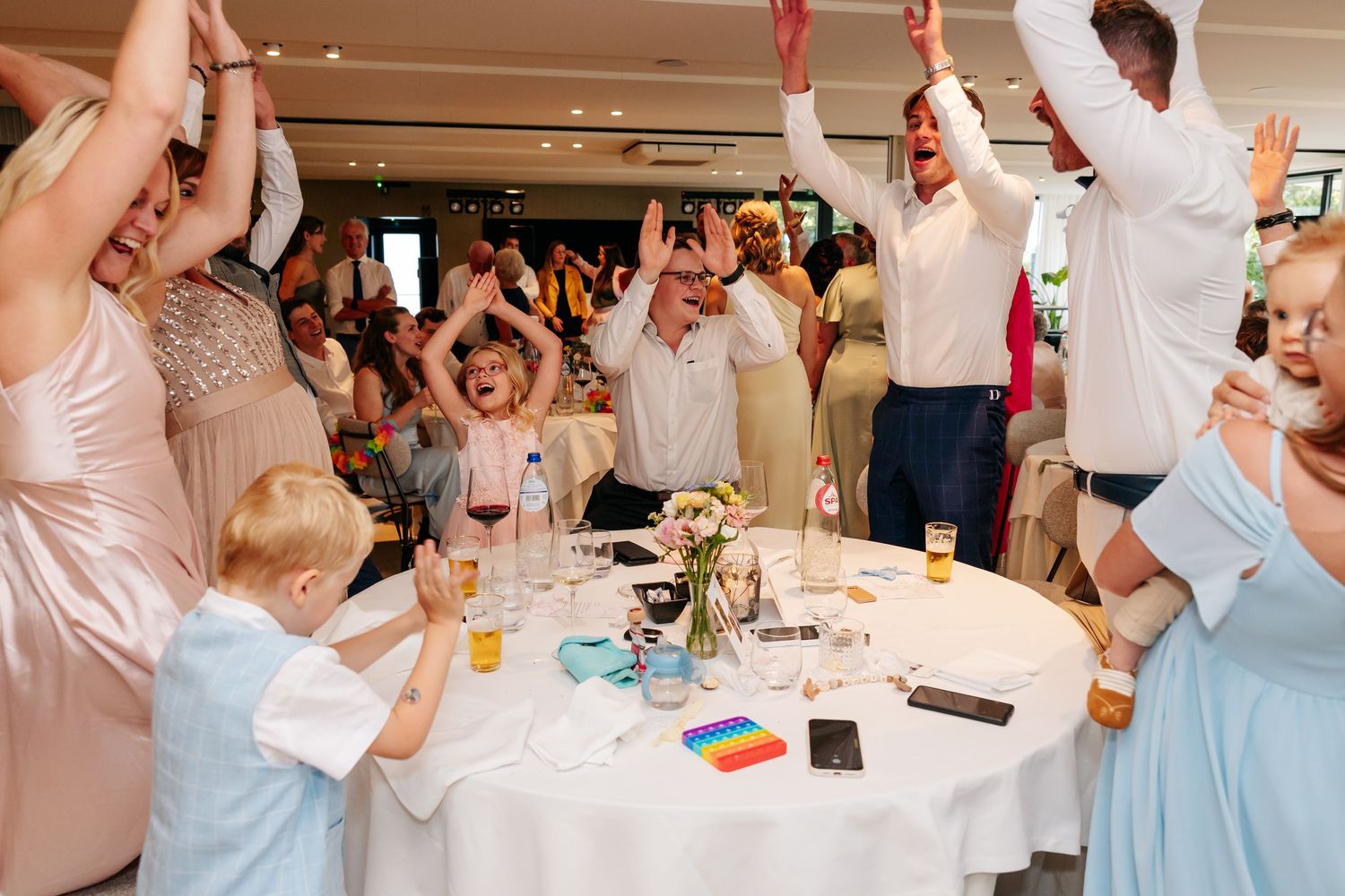 Joyful wedding guests and children dance with arms raised at reception.