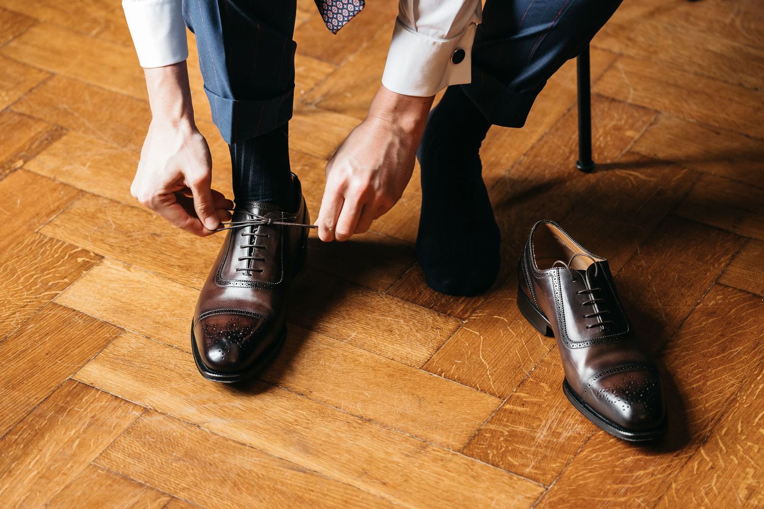 Groom tying brown leather Oxford brogue shoes on a parquet wood floor.
