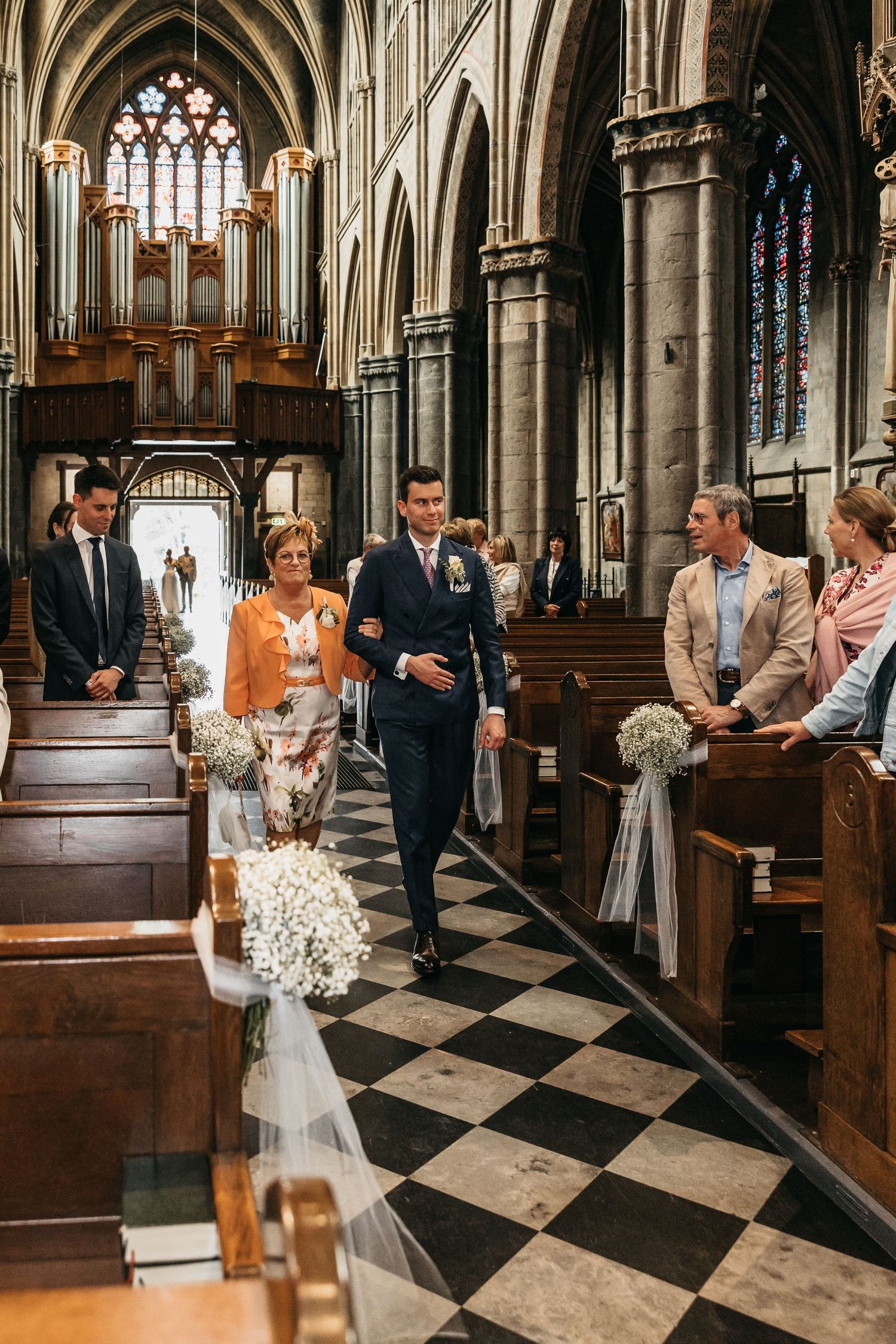 Groom walks down church aisle with mother during wedding ceremony in grand Gothic cathedral with checkered floors.