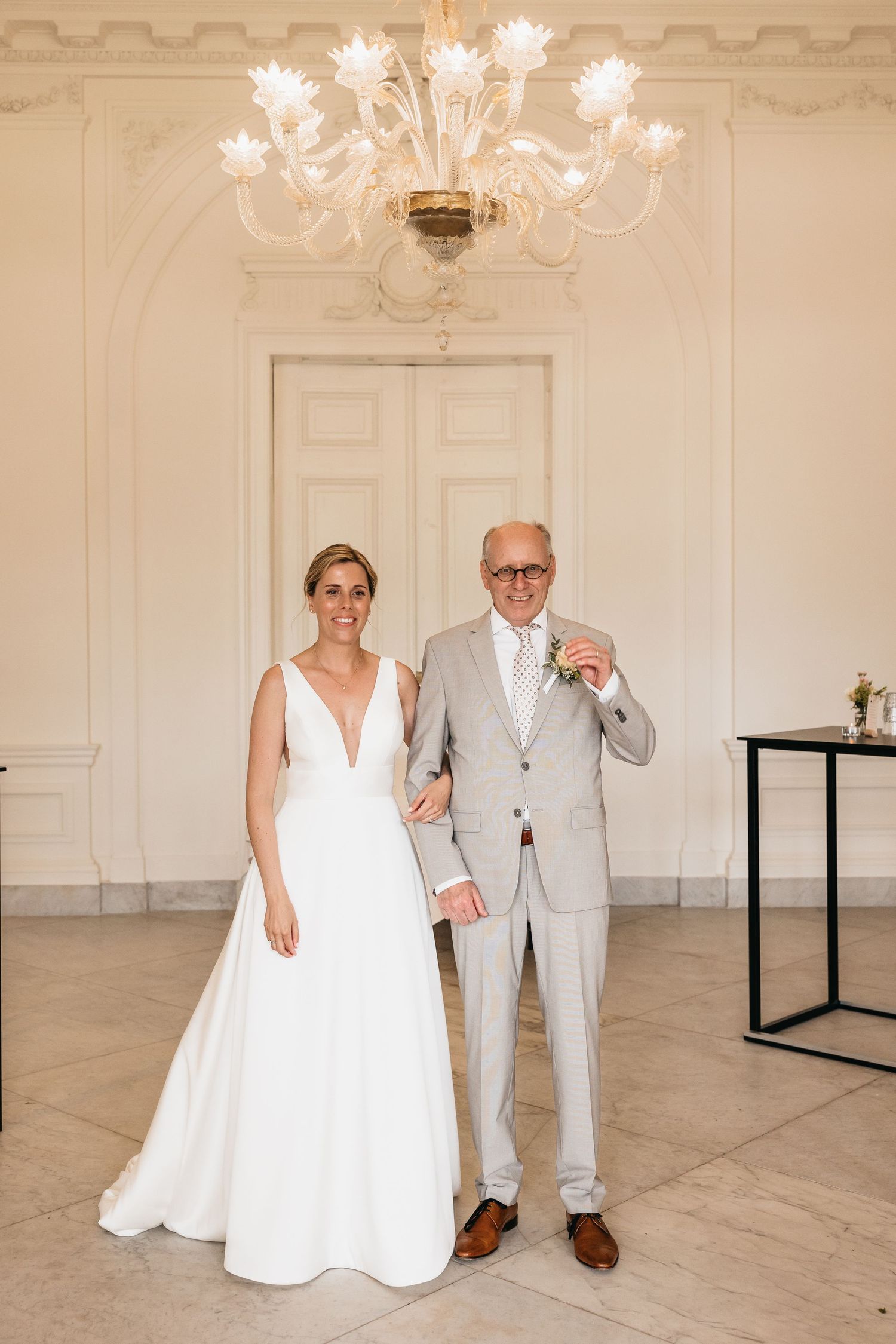 Smiling bride in deep V-neck white gown stands with older gentleman in grey suit under ornate chandelier.