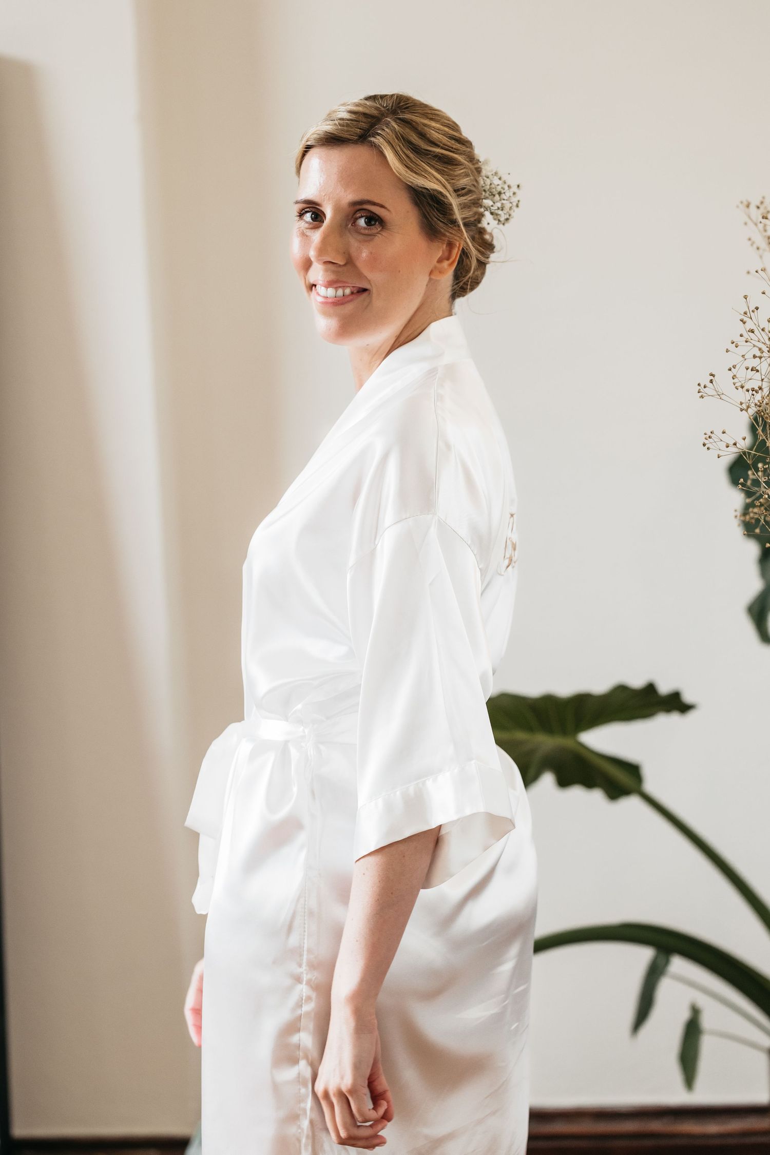 Smiling bride in a white satin robe with floral hair accessory standing in a bright room.