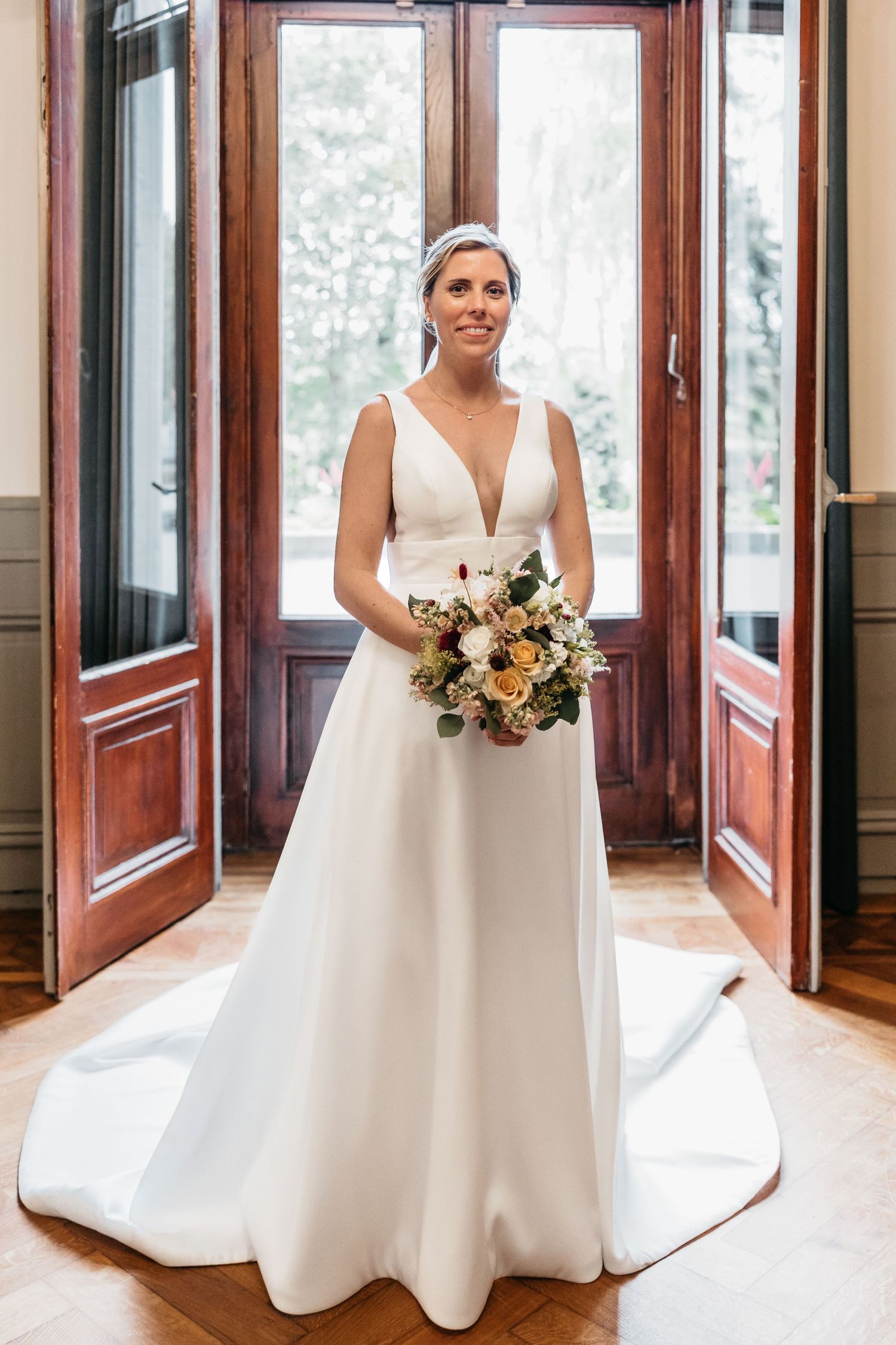 Bride in elegant deep V-neck white gown with train holding bouquet, standing before ornate wooden doors.