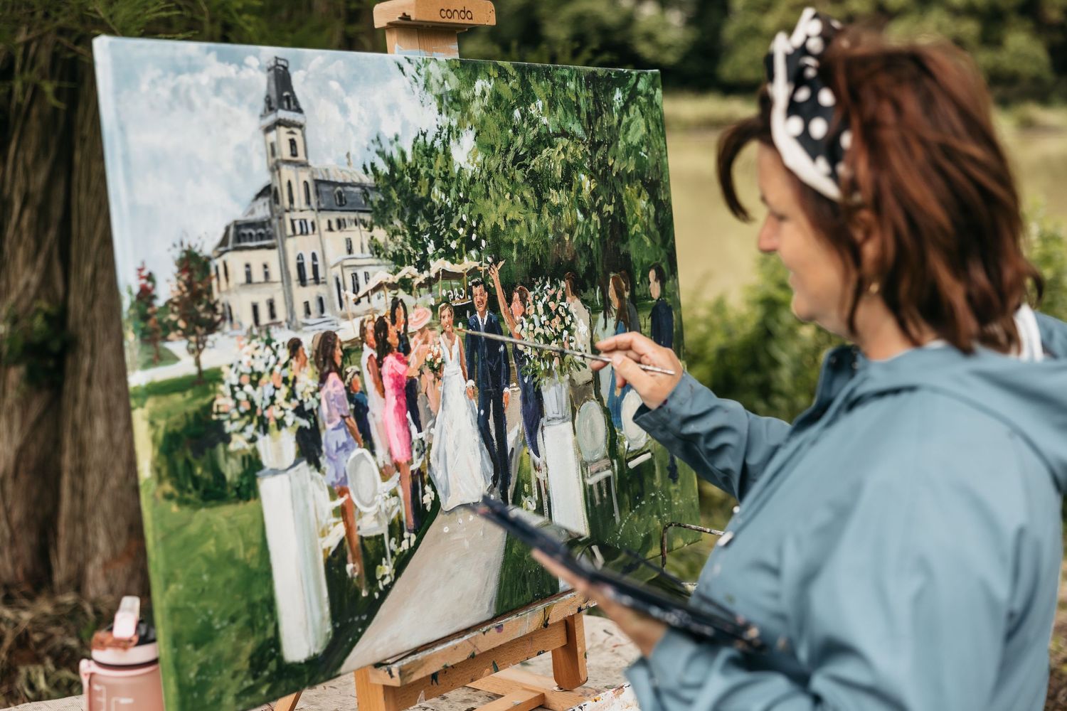 Artist painting live wedding scene on canvas at outdoor ceremony near historic church.