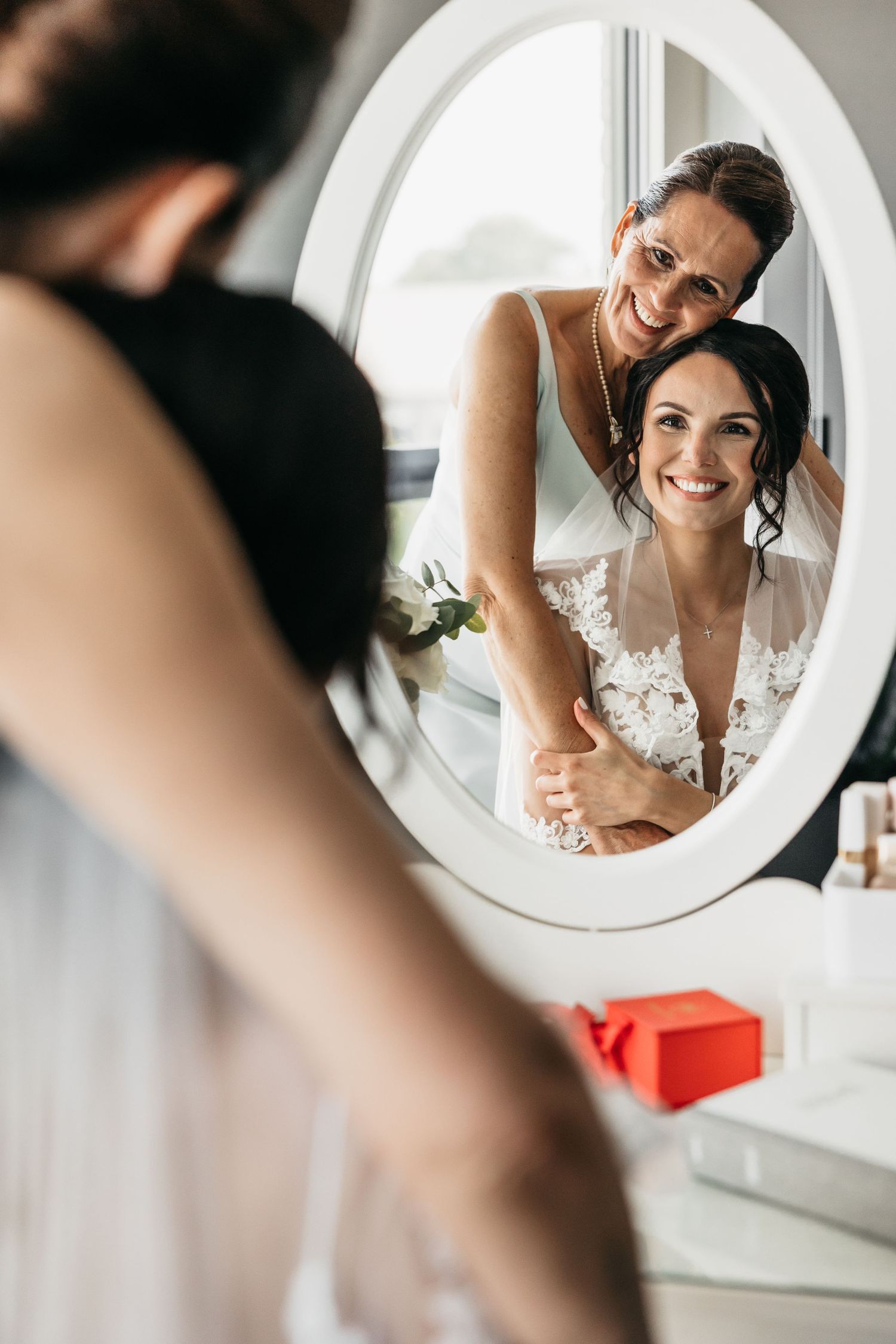 Joyful bride and bridesmaid smiling together in oval vanity mirror reflection during wedding preparations.