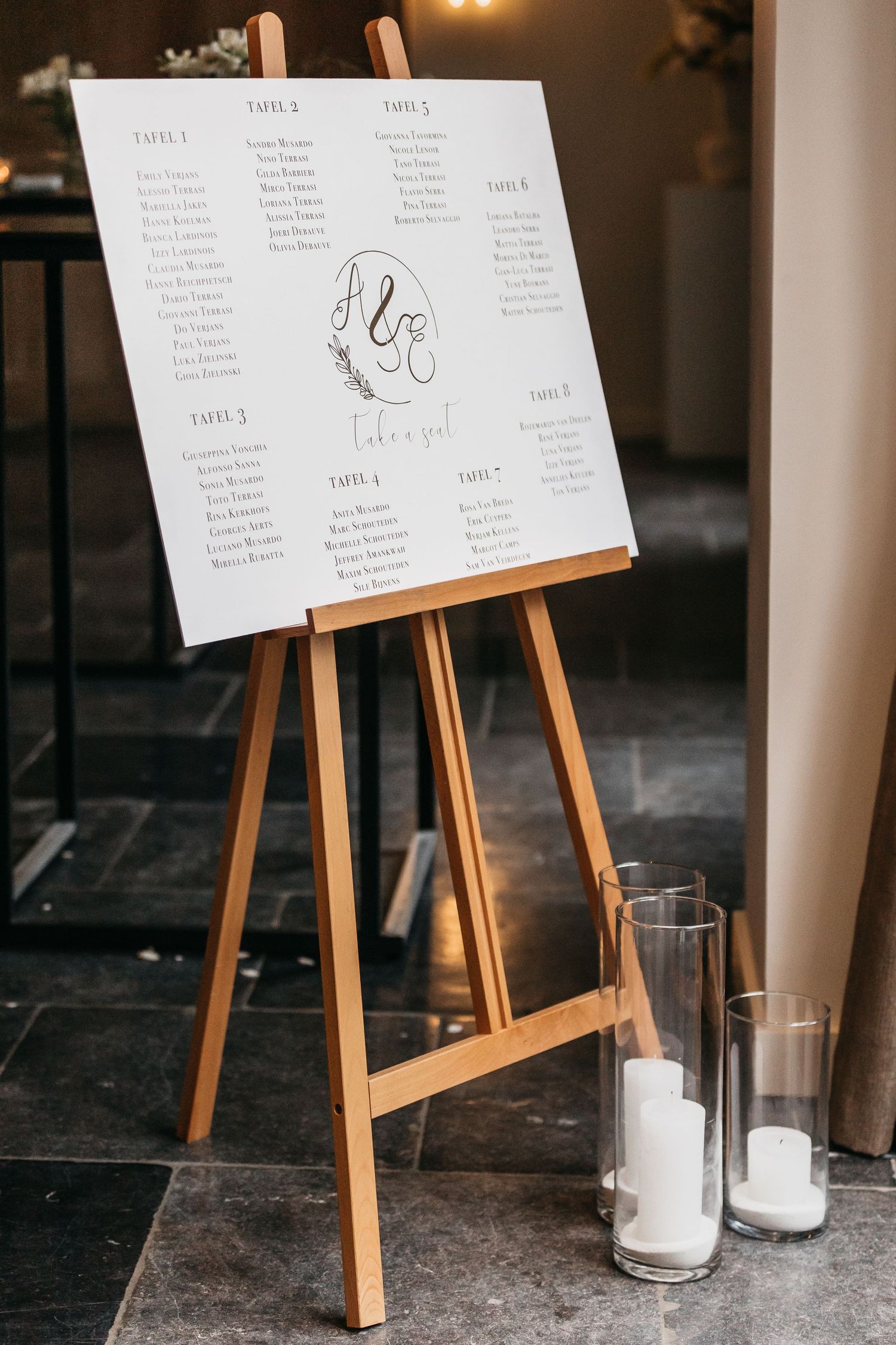 Wedding seating chart displayed on white board atop wooden easel with candles on dark tile floor.