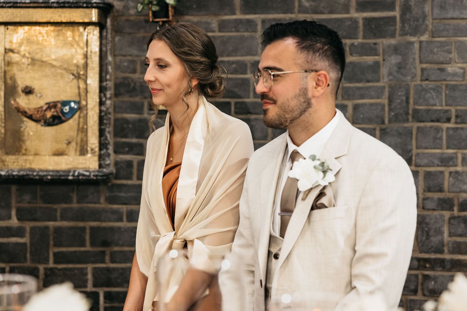 Elegant couple in cream and rust outfits seated together at wedding reception table.