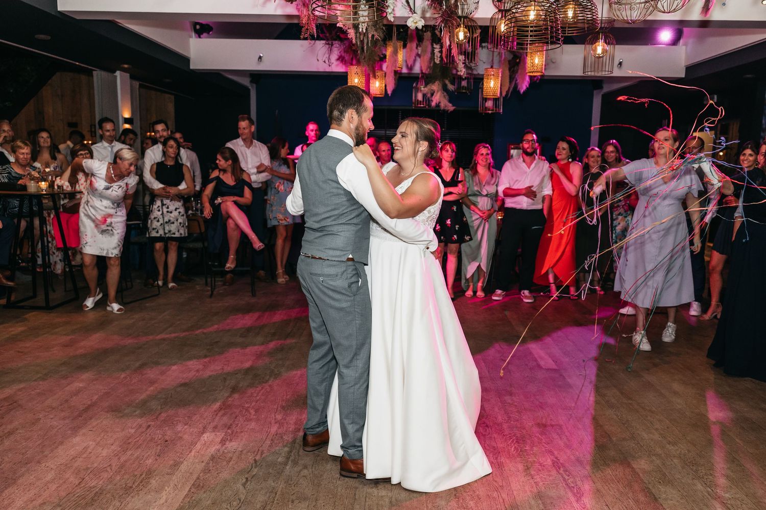 Bride in white gown and groom in grey suit share their first dance as guests watch at an elegant wedding reception.