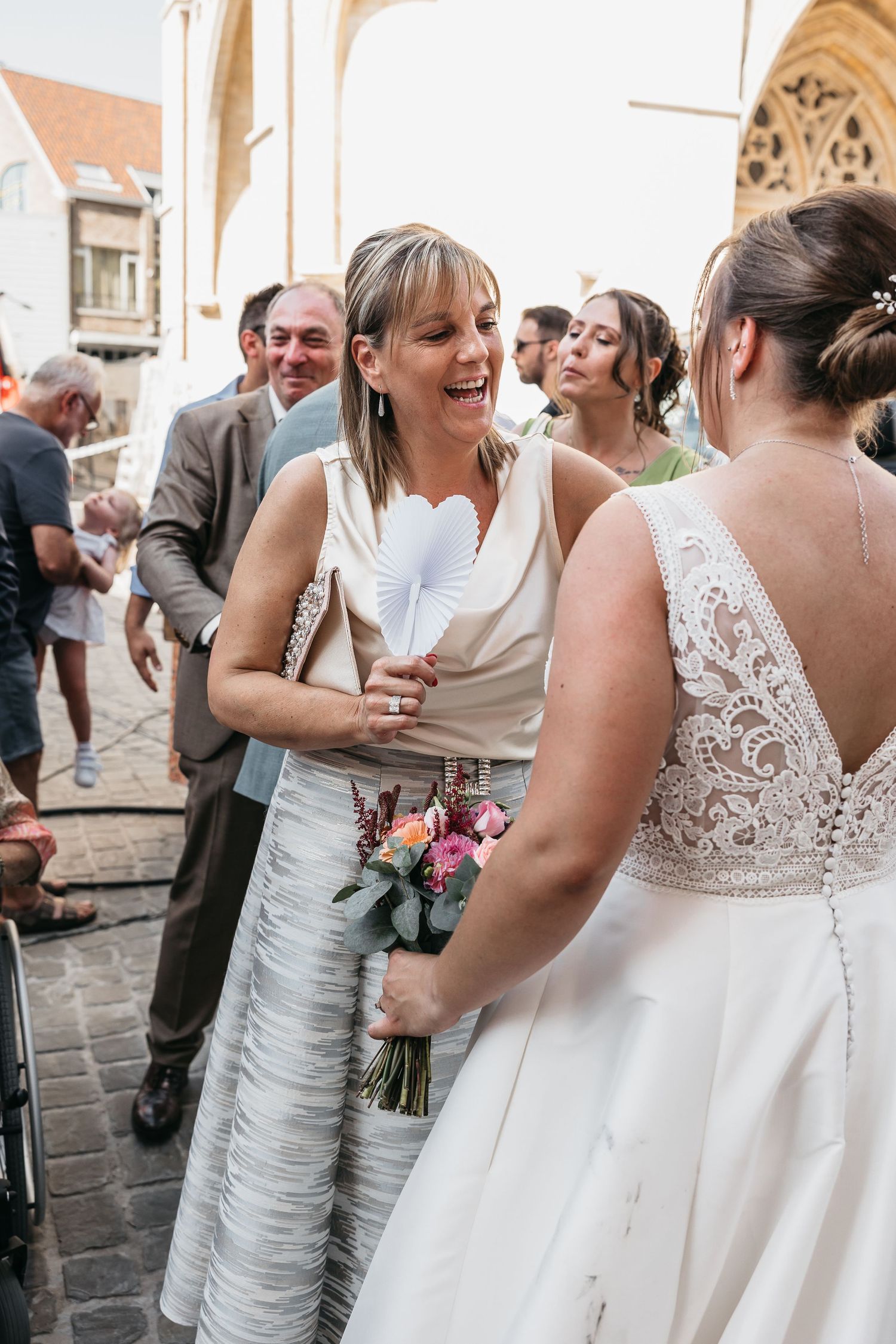 Joyful woman in white dress laughing with bride holding colorful bouquet at outdoor wedding reception.