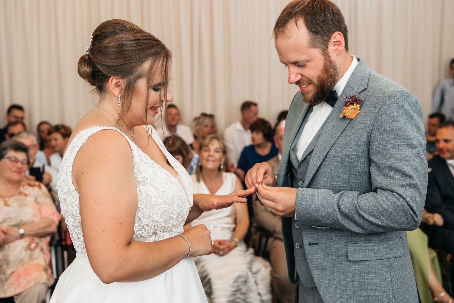 Smiling bride places a wedding ring on the grooms finger during their ceremony with guests watching.