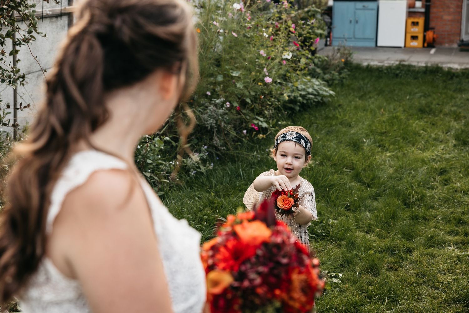 Young flower girl smiling and offering a red flower to the bride holding a colorful bouquet.