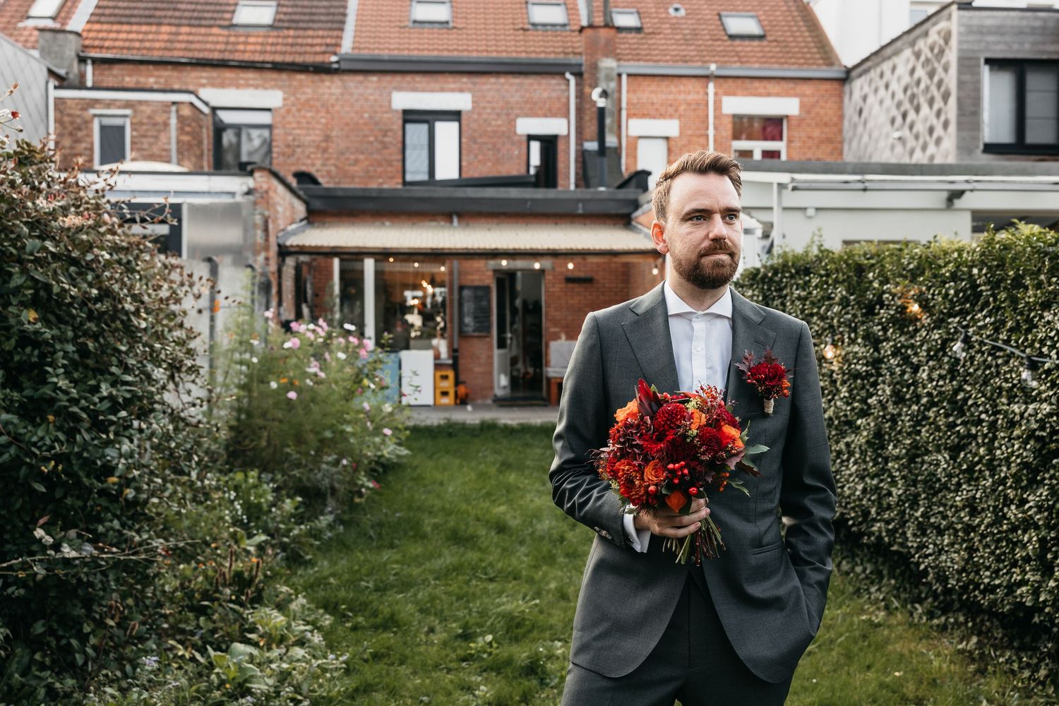 Groom in grey suit holding a vibrant red and orange bridal bouquet in a garden setting.