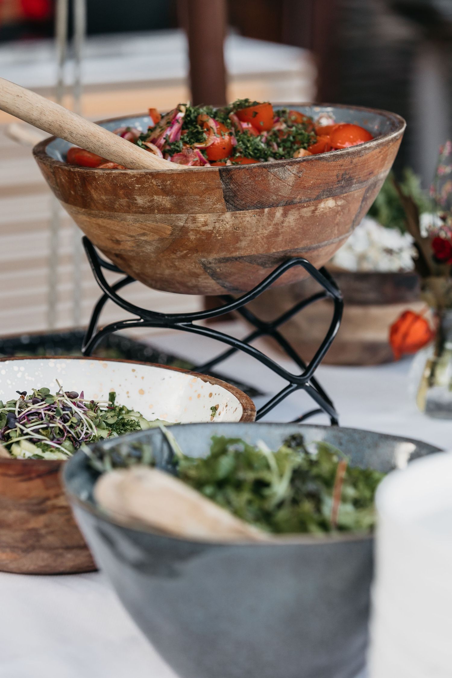 Wooden bowls of fresh tomato salad and green salads displayed on a white outdoor buffet table.