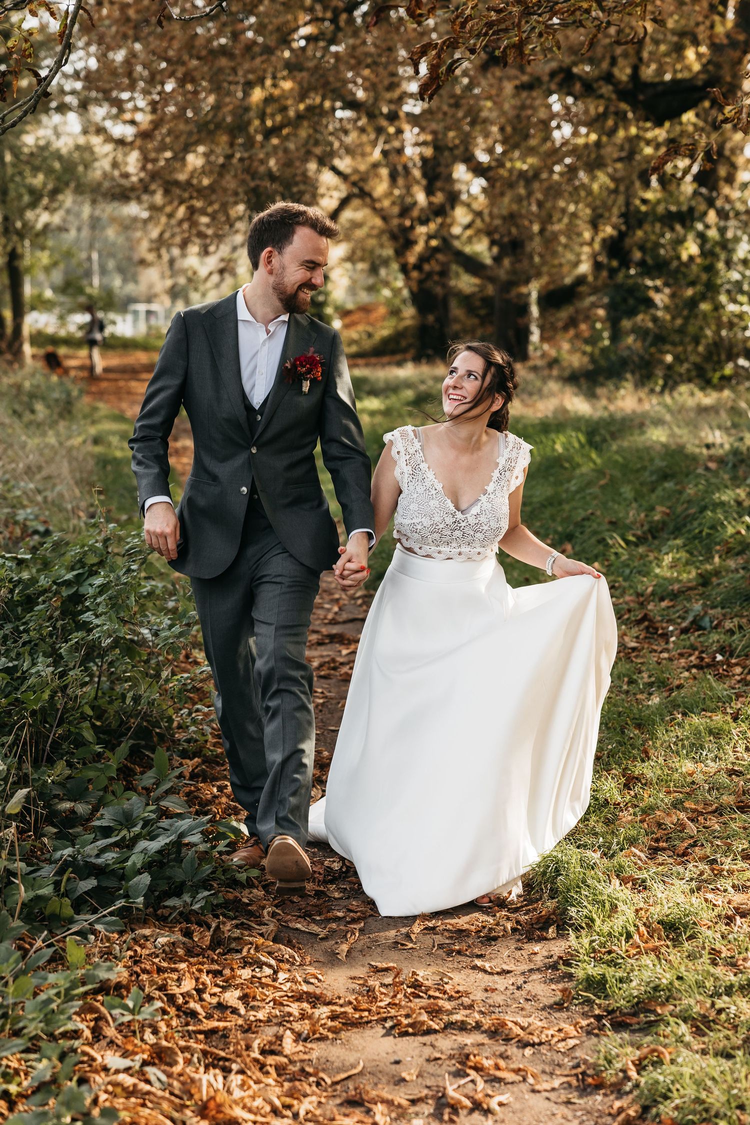 Laughing bride and groom walking hand in hand through a colorful autumn park on their wedding day.