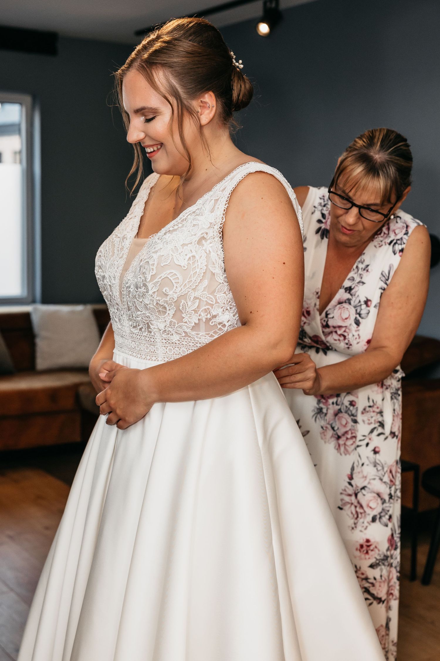 Bride smiling as woman in floral dress buttons up her lace and satin A-line wedding gown indoors.