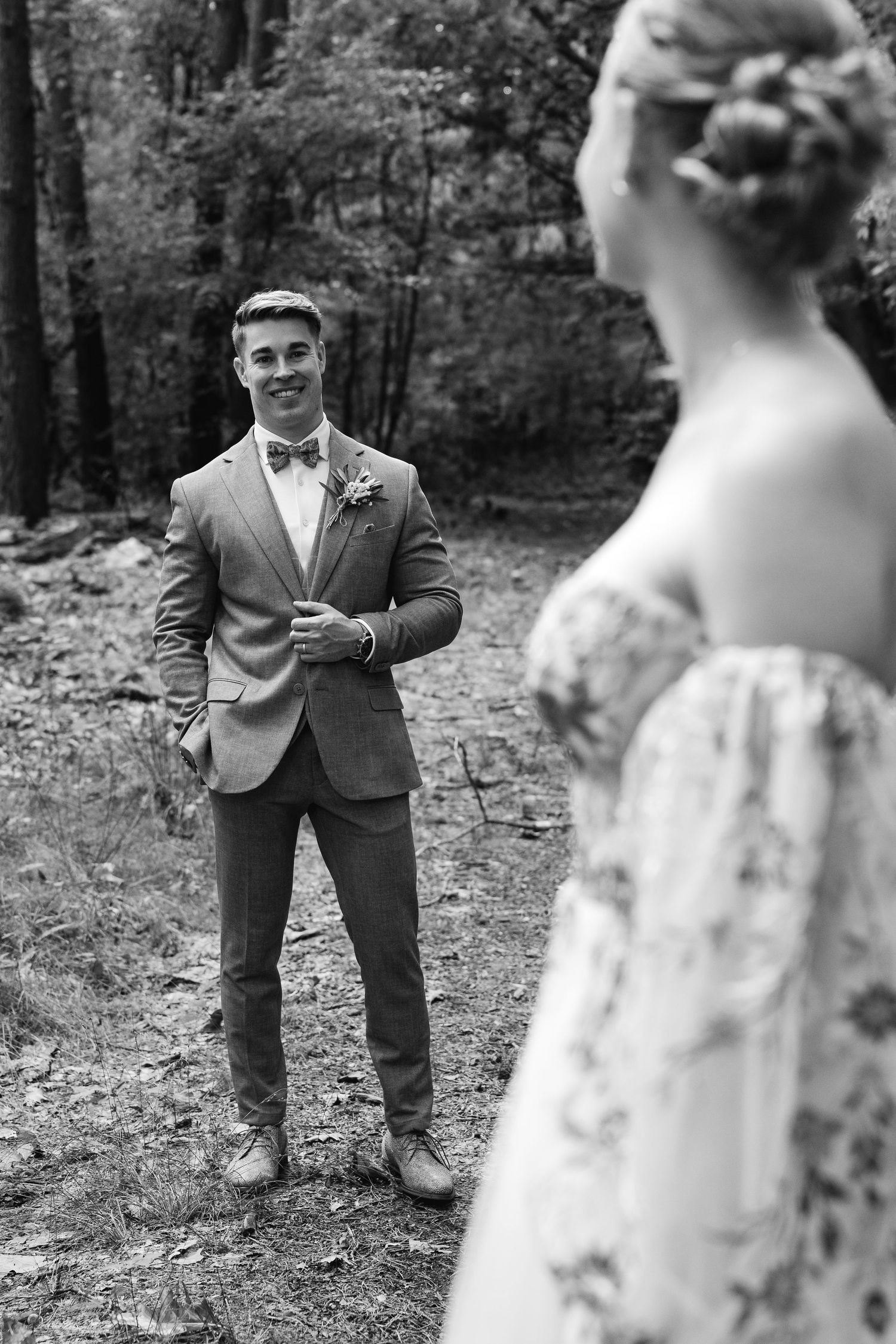 Black and white photo of smiling groom in suit with bow tie looking at bride in forest.