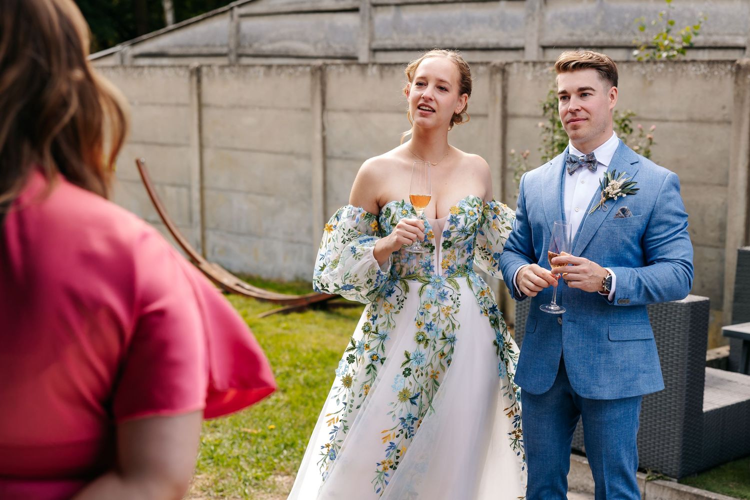 Bride in floral gown and groom in blue suit chat with guest at outdoor wedding.