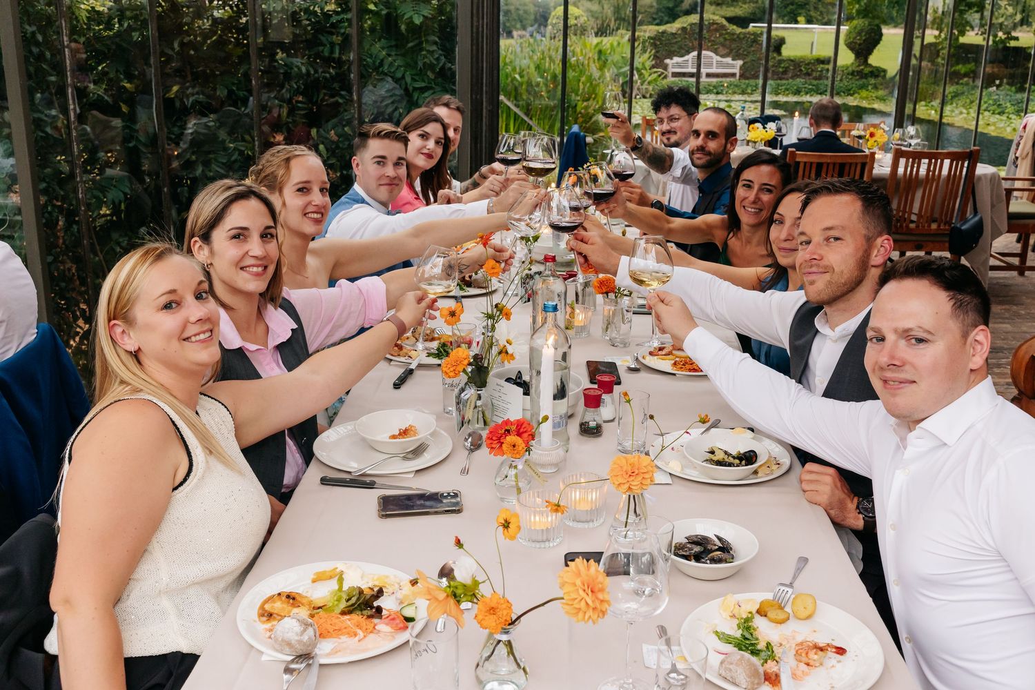Group of wedding guests raising glasses in a toast at an elegant outdoor dining table with floral centerpieces.