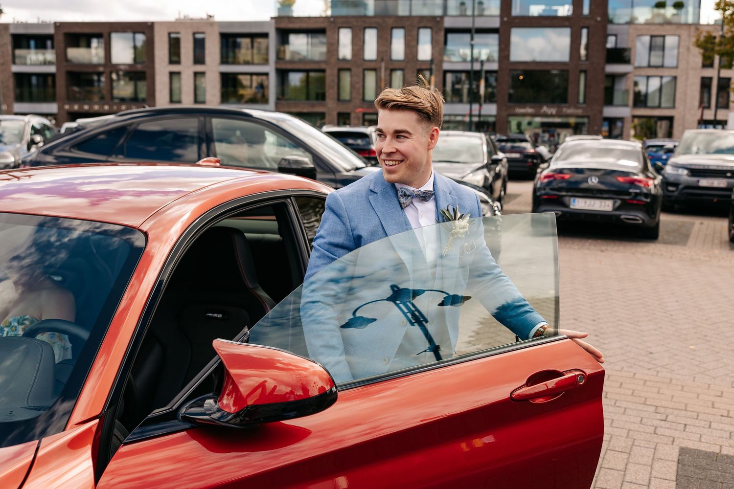 Smiling groom in blue suit opening door of red sports car at wedding.